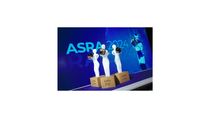 Das Bild zeigt drei Trophäen des ASRA 2024 (Austrian Sustainability Reporting Award), die auf einer Bühne präsentiert werden. Die stilisierten weißen Figuren mit einer bunten Kugel als Symbol halten ihre Arme nach oben und stehen auf Holzsockeln mit der Gravur „ASRA 2024“. Im Hintergrund ist eine große LED-Wand mit dem Schriftzug „ASRA 2024“ zu sehen, die das Event visuell unterstreicht. Der ASRA zeichnet Unternehmen für exzellente Berichterstattung und besonderes Engagement im Bereich Nachhaltigkeit und Corporate Social Responsibility (CSR) aus