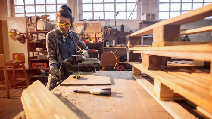 Eine Frau arbeitet in einer Werkstatt mit Holz. Sie trägt eine Schutzbrille und Gehörschutz.