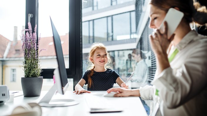 Arbeitssituation mit Kind Frau sitzt am Schreibtisch und telefoniert, daneben steht ein kleines Mädchen und sieht ihr zu