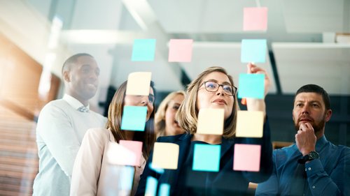 Das Bild zeigt eine Gruppe von Menschen vor einer Glaswand, die mit bunten Haftnotizen bedeckt ist. Die Personen scheinen in einer Besprechung oder Brainstorming-Sitzung zu sein und schauen auf die Notizen, während sie diskutieren und Ideen austauschen. Die Haftnotizen sind in verschiedenen Farben wie Blau, Gelb und Rosa. Die Szene findet in einem modernen Büro oder Konferenzraum mit heller Beleuchtung statt.