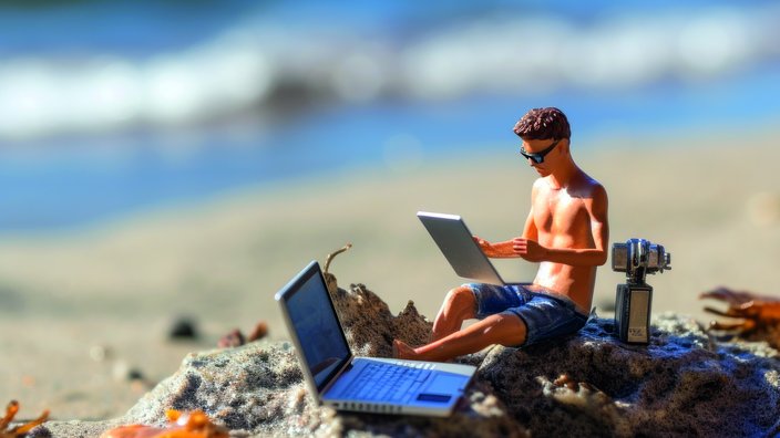 Miniaturfigur eines Mannes mit Sonnenbrille, der oberkörperfrei und in Shorts an einem Strand sitzt und an einem Laptop arbeitet. Um ihn herum befinden sich weitere technische Geräte, darunter ein zweiter Laptop und eine Kamera