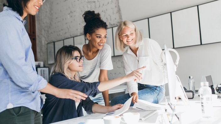 Das Bild zeigt vier Frauen sitzend und stehend um einen Schreibtisch. Alle vier Frauen schauen auf den Bildschirm.