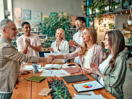 Das Bild zeigt eine Gruppe von sechs Personen in einem modernen, grünen Büro applaudiert, während ein älterer Mann und eine jüngere Frau sich lächelnd die Hand geben. Auf dem Holztisch vor ihnen liegen Unterlagen, Laptops und Diagramme.