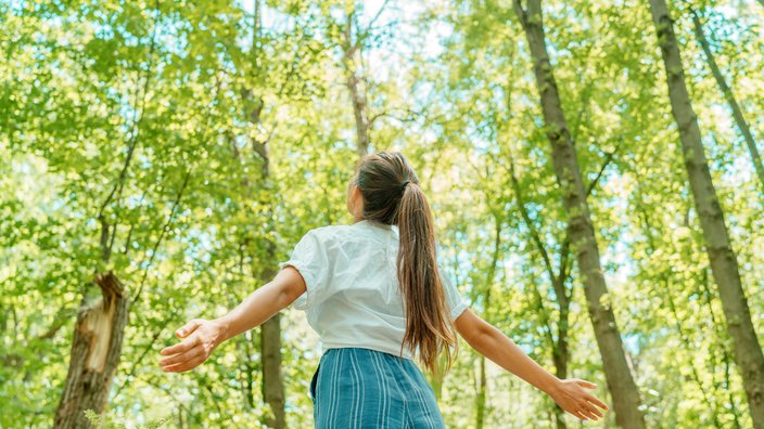 Das Bild zeigt eine junge Frau, die im Wald steht und die Arme hebt.