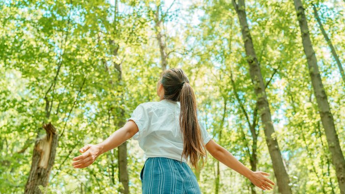 Junge Frau steht im Wald Das Bild zeigt eine junge Frau, die im Wald steht und die Arme hebt.