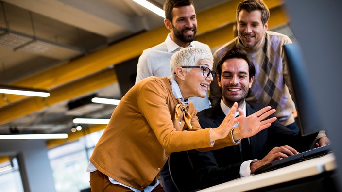 Generationenübergreifendes Arbeiten im Team Das Bild zeigt eine ältere Frau, die drei Männern vor einem Bildschirm etwas erklärt.