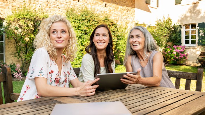 Drei Frauen sitzen an einem Tisch im Garten und schauen lachend in die Ferne. Sie halten ein Tablet in der Hand.