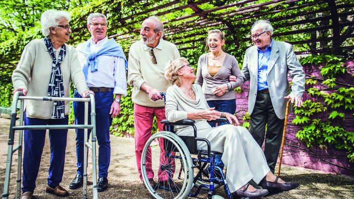Das Bild zeigt eine Gruppe Senior:innen in einer Laube stehend. Sie sind Bewohner:innen eines Pflegeheims. Eine Person sitzt im Rollstuhl, eine Person benutzt einen Rollator.