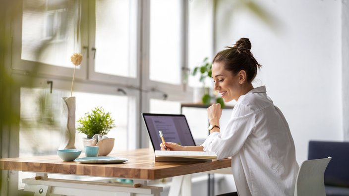 Das Bild zeigt eine junge Frau an ihrem Schreibtisch sitzend. Vor ihr steht ein Laptop und sie lächelt fröhlich.