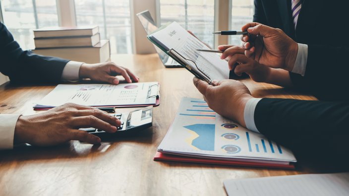 Drei Geschäftspersonen analysieren gemeinsam Finanzdokumente an einem Holztisch. Auf dem Tisch liegen Diagramme, Berichte, ein Taschenrechner und ein Laptop. Eine Person hält ein Klemmbrett mit einem Bericht, während die anderen mit Stift und Finger auf Daten zeigen. Im Hintergrund sind Bücher und große Fenster zu sehen.
