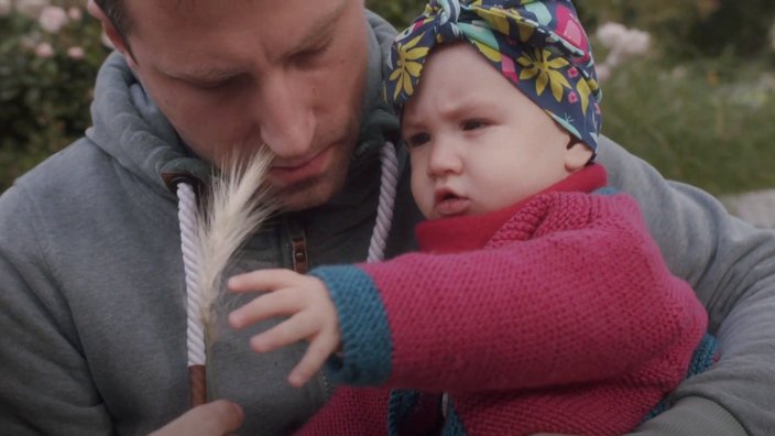 Ein Mann hält ein Baby im Arm. Beide betrachten neugierig eine Ähre, die der Mann in der Hand hält. Das Baby trägt eine bunte Kopfbedeckung und eine pinke Strickjacke, der Mann einen grauen Hoodie