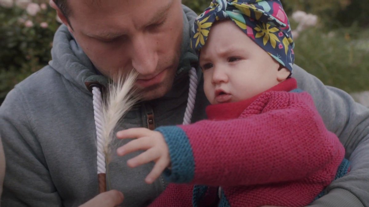 Ein Mann hält ein Baby im Arm. Beide betrachten neugierig eine Ähre, die der Mann in der Hand hält. Das Baby trägt eine bunte Kopfbedeckung und eine pinke Strickjacke, der Mann einen grauen Hoodie