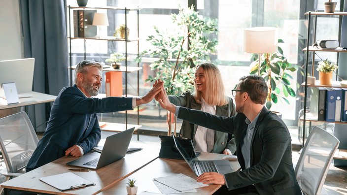 Das Bild zeigt zwei Männer und eine Frau die sich in einem modernen Büro gegenüber sitzen und sich lachend ein High Five geben