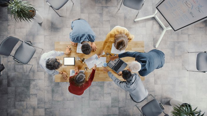 Das Bild zeigt sechs Personen aus der Vogelperspektive an einem Tisch sitzend. 