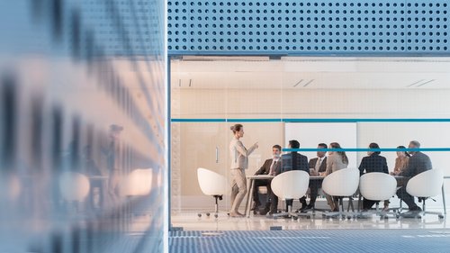 Ein moderner Konferenzraum mit Glaswänden, in dem ein Geschäftstreffen stattfindet. Acht Personen sitzen um einen Tisch, und eine stehende Person präsentiert etwas vor der Gruppe. Der Raum hat ein sauberes, professionelles Erscheinungsbild mit weißen Stühlen und einem blauen Streifen, der horizontal über die Glaswände verläuft.