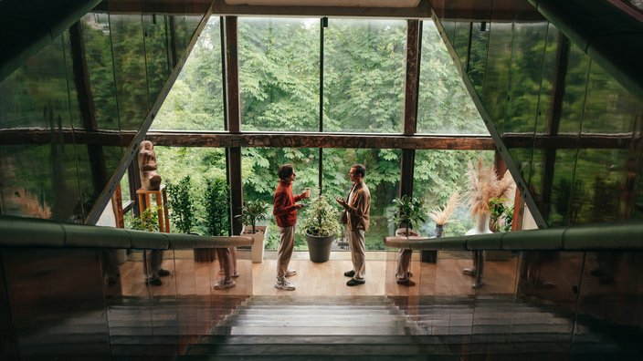 Das Bild zeigt zwei Personen, die sich auf einem Treppenabsatz eines modernen Gebäudes unterhalten. Sie stehen vor einer großen Fensterfront mit Blick auf dichtes, grünes Blattwerk von Bäumen draußen.