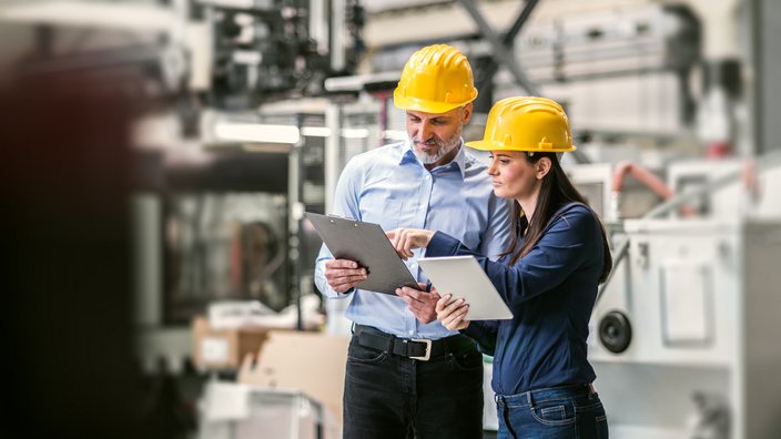 Ein Mann und eine Frau stehen in einer Industrieanlage. Sie tragen Schutzhelme und sehen auf ihre Klemmbretter.