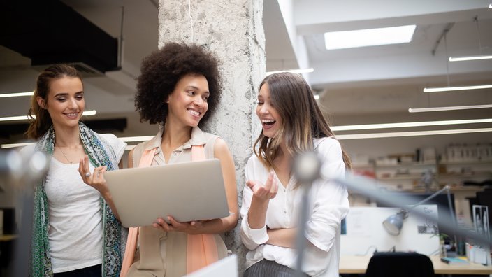 Teamwork und gute Laune im modernen Büro Das Bild zeigt eine Besprechung von drei Frauen stehend mit einem Laptop in der Hand.