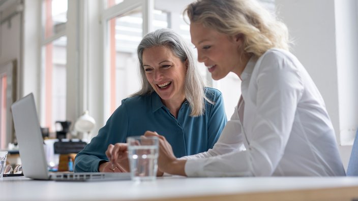Das Bild zeigt zwei Frauen an einem Schreibtisch und vor einem Laptop sitzend. Die beiden Frauen unterhalten sich fröhlich und blicken lächelnd auf den Laptop.