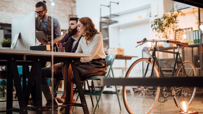 Teamwork in moderner Arbeitsumgebung Das Bild zeigt drei Personen in einem modernen Büro, die sich um einen Schreibtisch mit einem Computerbildschirm und verschiedenen Büromaterialien versammelt haben. Eine Person steht und zeigt den anderen beiden, die sitzen, etwas auf einem Tablet. Im Hintergrund sind ein Fahrrad, Bücherregale und Pflanzen zu sehen. Das Bild vermittelt eine kollaborative Arbeitsatmosphäre.