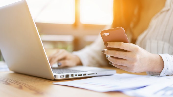Ds Bild zeigt eine Frau, die beim Laptop sitzt und arbeitet. In ihrer linken Hand hält sie zusätzlich ein Smartphone.