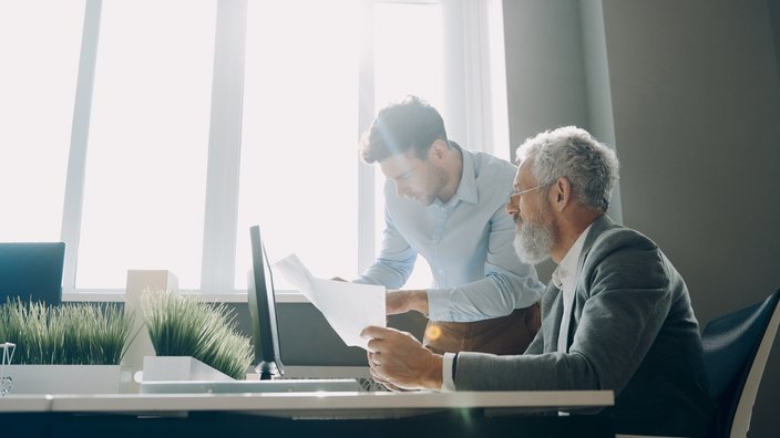Teamwork in moderner Arbeitsumgebung Das Bild zeigt eine Gesprächssituation zwischen zwei Männern. Der ältere Herr sitz an einem Tisch während der jüngere Mann neben ihm steht.