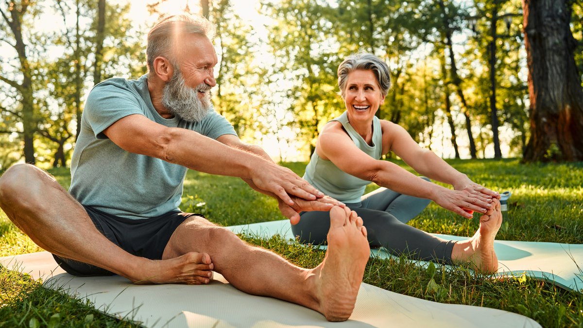 Das Bild zeigt ein älteres Ehepaar auf Yogamatten sitzend, bei Dehnungsübungen.