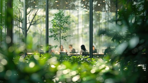 Das Bild zeigt einen üppigen, grünen Innenraumgarten mit verschiedenen Pflanzen und Bäumen im Vordergrund. Durch ein großes Fenster im Hintergrund sind mehrere Personen zu sehen, die an einem Tisch sitzen, möglicherweise in einem Meeting oder bei der Arbeit. Der Fokus liegt auf der Pflanzenvielfalt im Vordergrund, während die Menschen und der Innenraum leicht unscharf dargestellt sind, was eine ruhige und natürliche Atmosphäre im Innenbereich erzeugt.