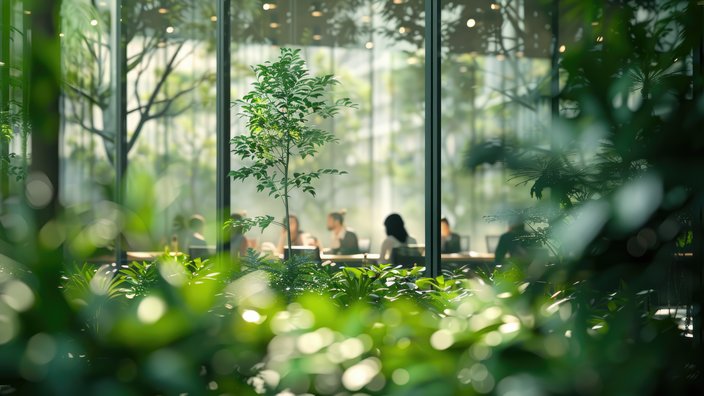 Grüner Arbeitsort Das Bild zeigt einen üppigen, grünen Innenraumgarten mit verschiedenen Pflanzen und Bäumen im Vordergrund. Durch ein großes Fenster im Hintergrund sind mehrere Personen zu sehen, die an einem Tisch sitzen, möglicherweise in einem Meeting oder bei der Arbeit. Der Fokus liegt auf der Pflanzenvielfalt im Vordergrund, während die Menschen und der Innenraum leicht unscharf dargestellt sind, was eine ruhige und natürliche Atmosphäre im Innenbereich erzeugt.