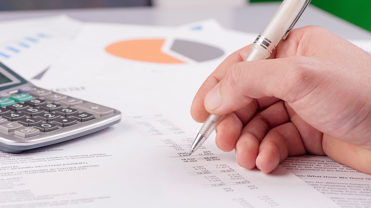Hand mit Kugelschreiber zeigt auf eine Finanzübersicht mit Zahlen und Tabellen; daneben liegt ein Taschenrechner, im Hintergrund ein Kreisdiagramm auf einem weiteren Blatt Papier.