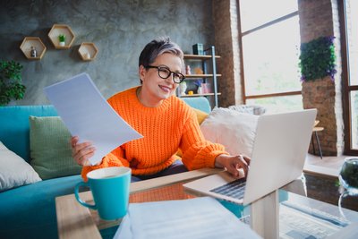 Das Bild zeigt eine Frau an einem Tisch sitzend. Vor ihr steht ein Laptop und ein Kaffeebecher. In ihrer rechten Hand hält sie Papierblätter.
